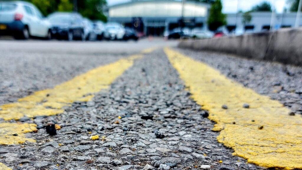 Close-up picture of double yellow lines on a road lined with cars on the opposite side